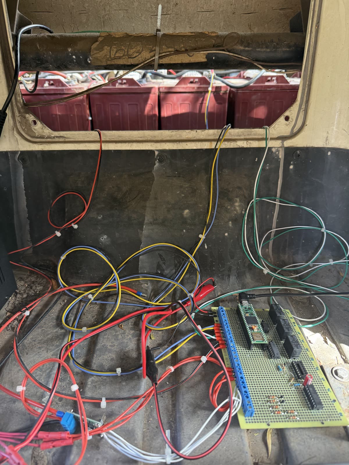 A soldered prototype board with a Teensy microcontroller, relays and terminal blocks, sitting on the cart's rear floor among zip-tied wiring, with the battery pack above